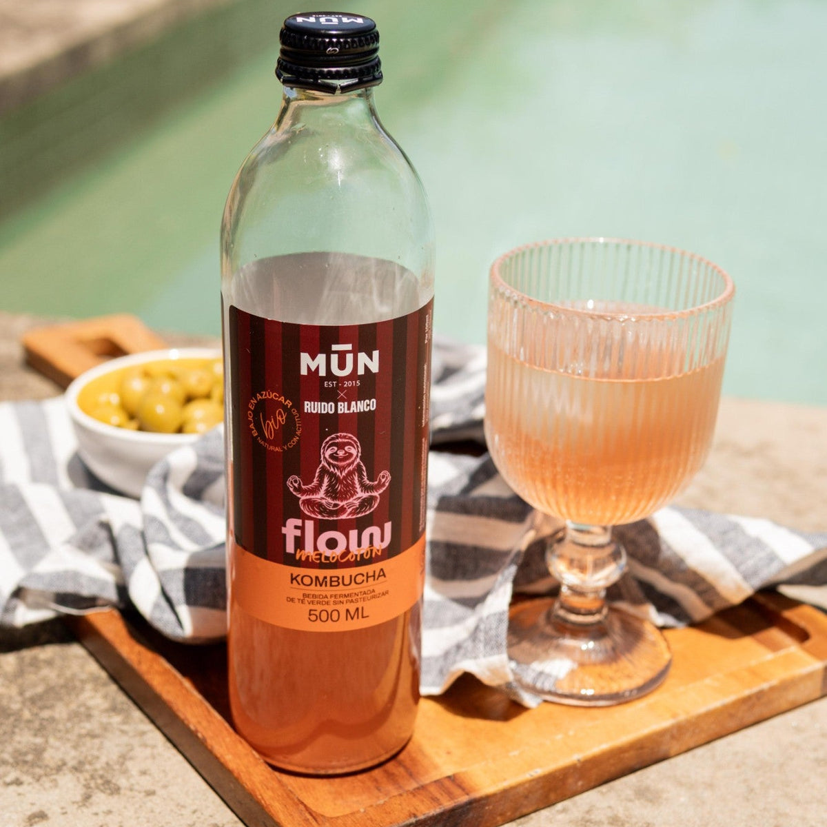 Botella de kombucha Mūn x Ruido Blanco FLOW melocotón de 500 ml junto a una copa al borde de la piscina